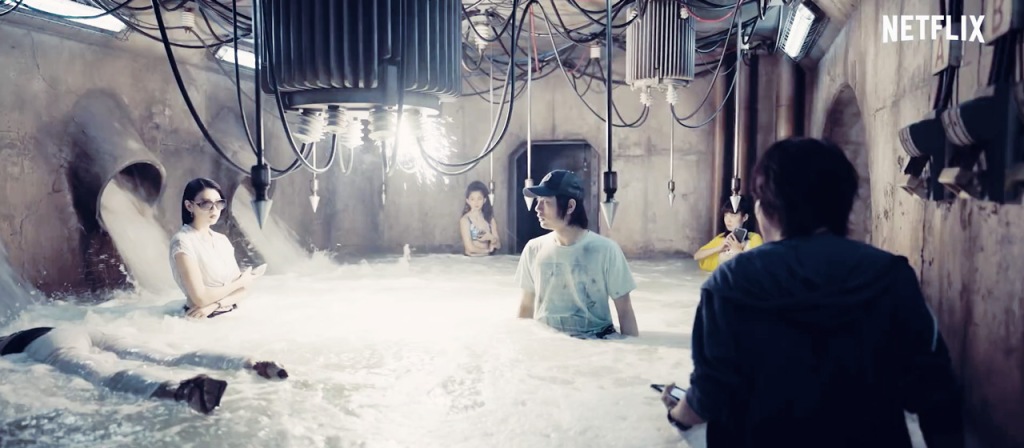 A screenshot from episode 5. A group of people standing in a room half-filled with water with electrical wires sparking 3 feet above. A dead body can be seen floating nearby.